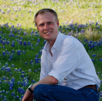 headshot of smiling man in a white shirt kneeling in a field of bluebonnets